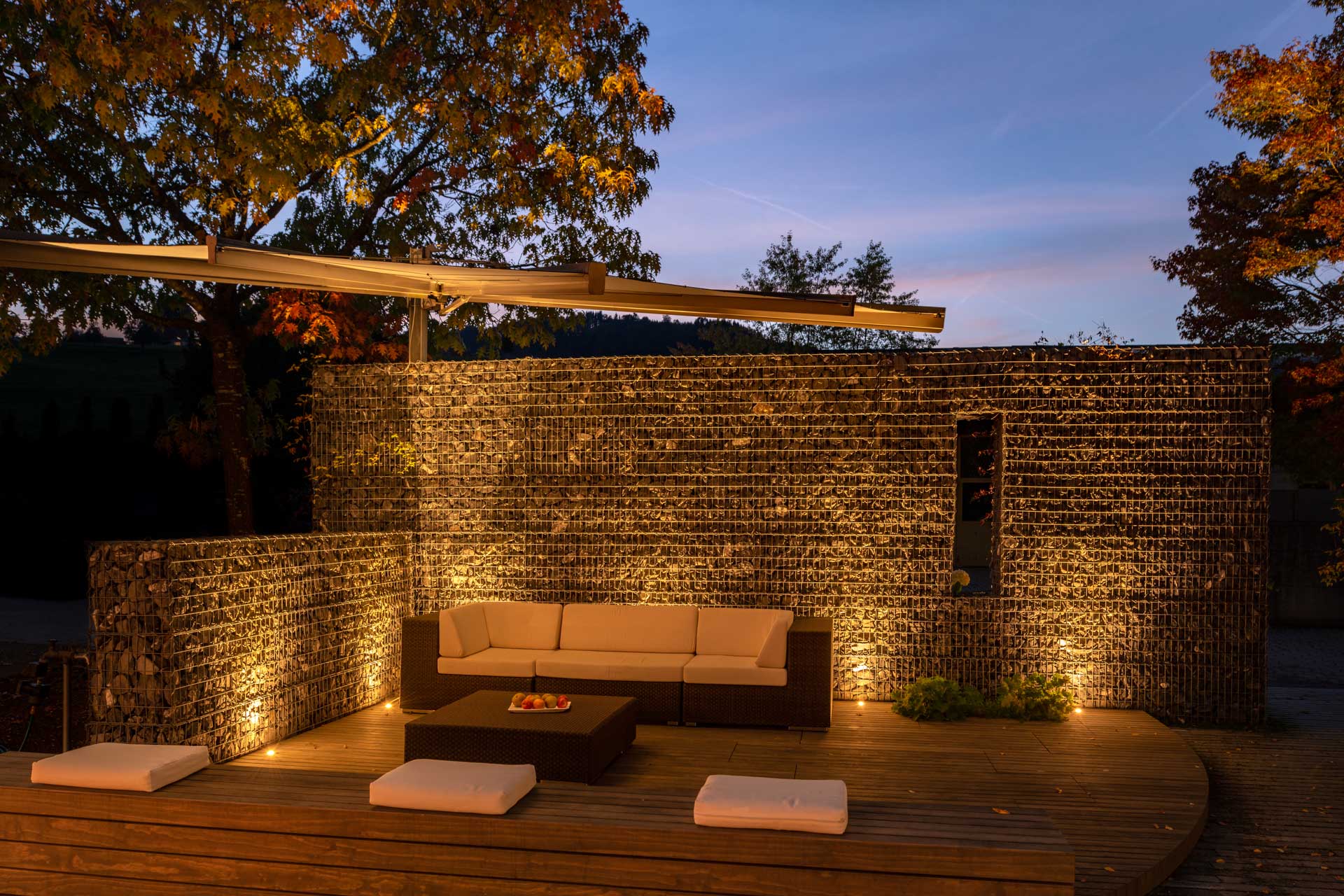 Steinmauer mit Superspot beleuchtet, hinter Terrasse mit Gartenmöbeln 